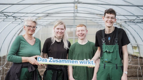 Vier Menschen im Gewächshaus halten ein Schild mit „Das Zusammen wirkt.“, Teamarbeit und Inklusion beim CJD.